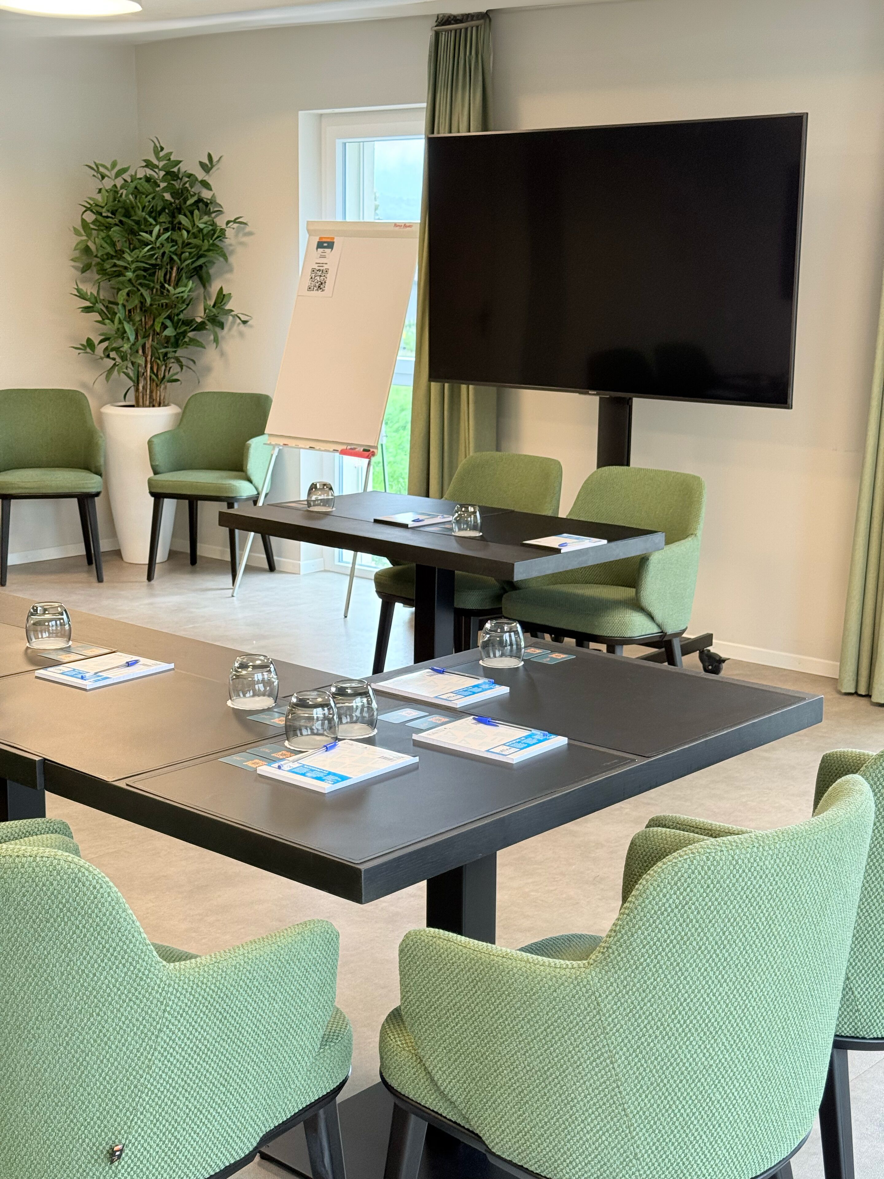 A conference room with a large table, green chairs, a flat-screen TV, a whiteboard, and a potted plant.