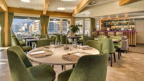 A restaurant with round tables, green upholstered chairs, a bar area with bottles, and a view of buildings through large windows. - Alparthotel  (Collombey-Muraz)