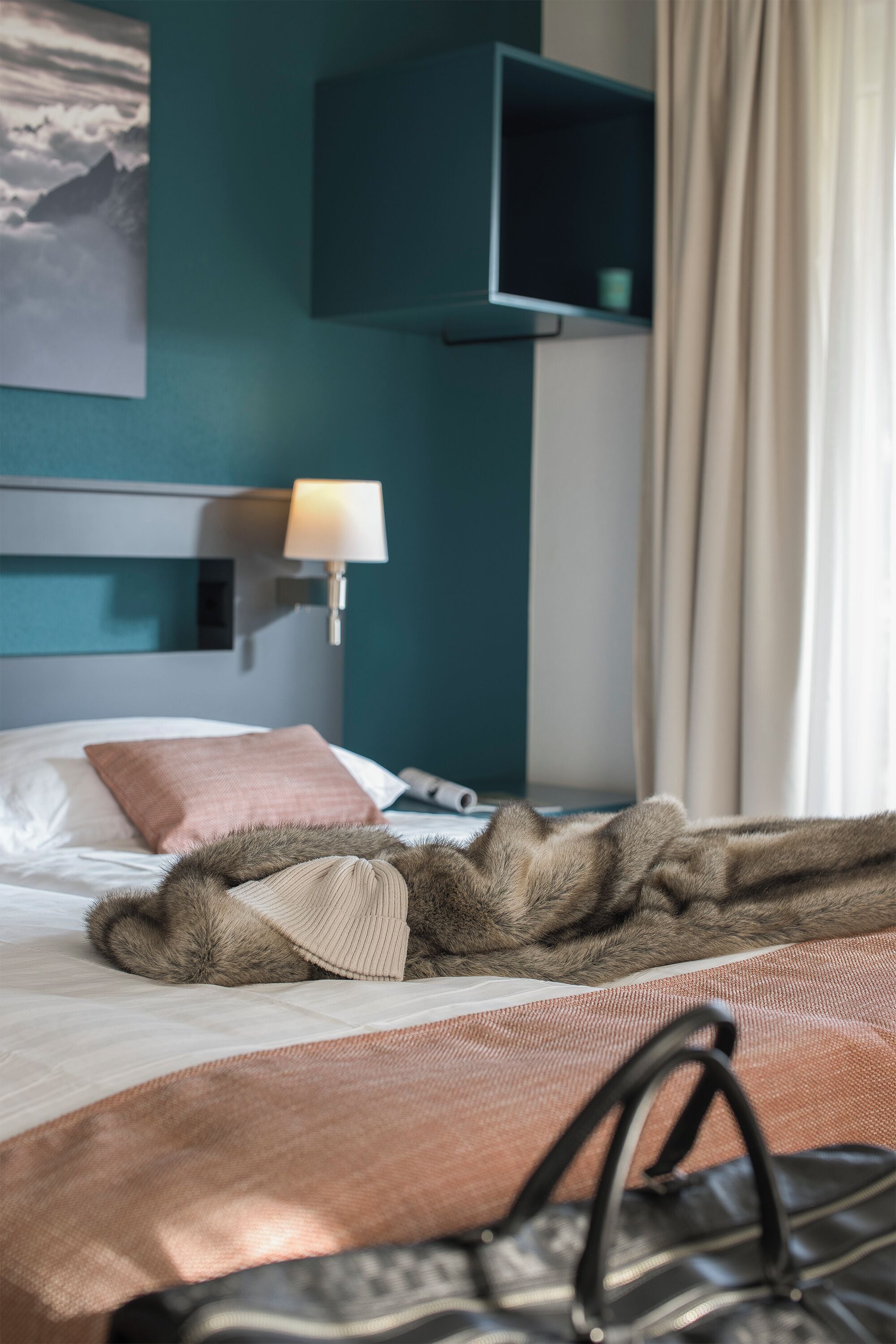 A hotel room with a bed, a lamp, a fur throw, and a handbag.