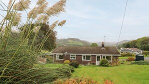 A single-story house with a brick exterior, a small front yard, and a view of rolling hills in the background. - Llwynon (Aberystwyth)