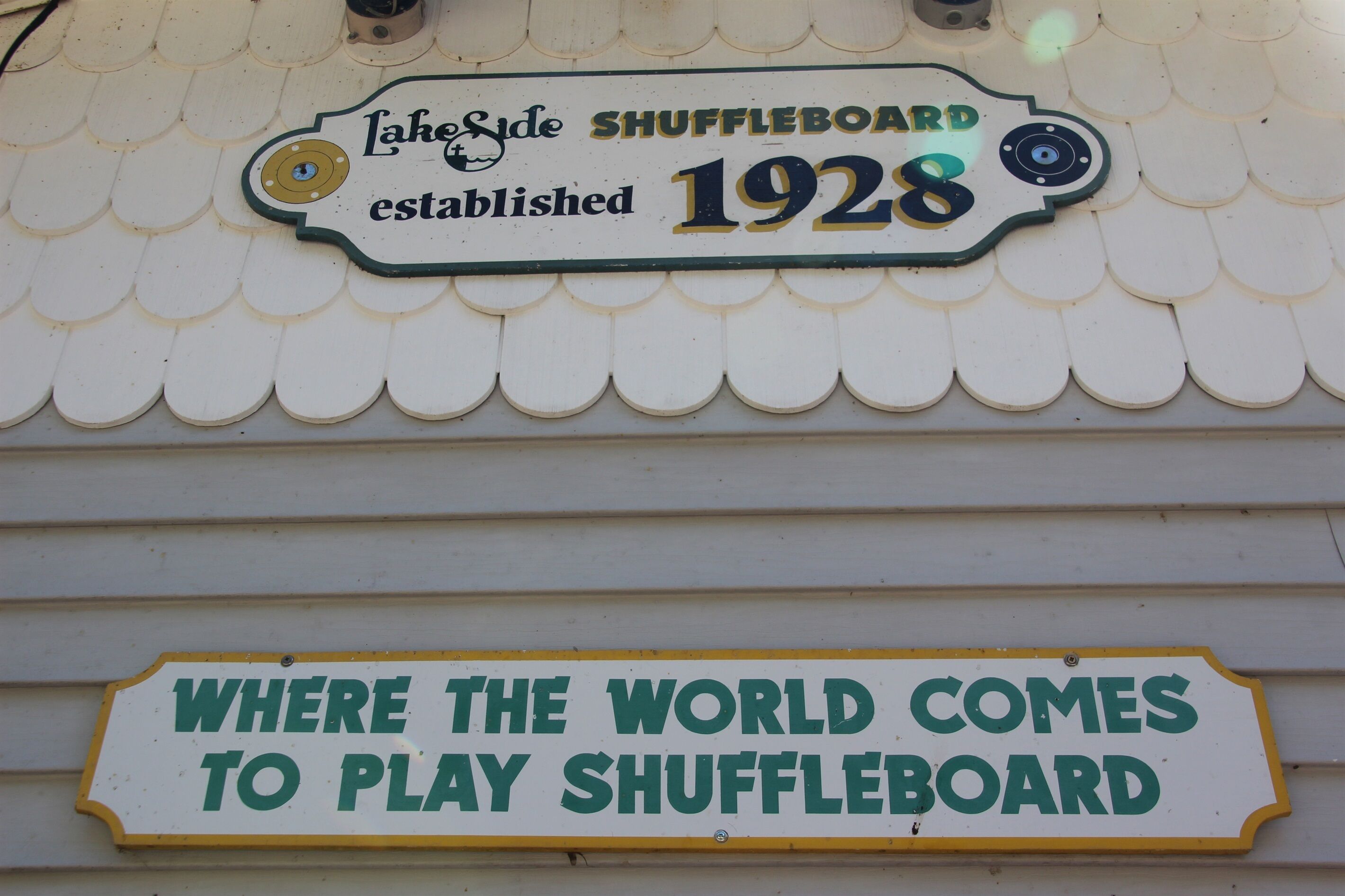 A sign with the text 'Lake Side Shuffleboard established 1928' and 'Where the world comes to play shuffleboard' on a building facade.