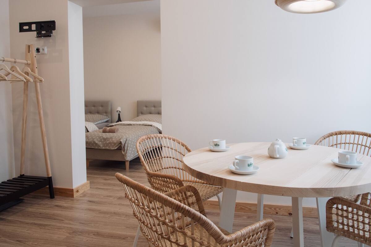 A modern, well-lit room with a round dining table, wicker chairs, and a bed with a headboard.