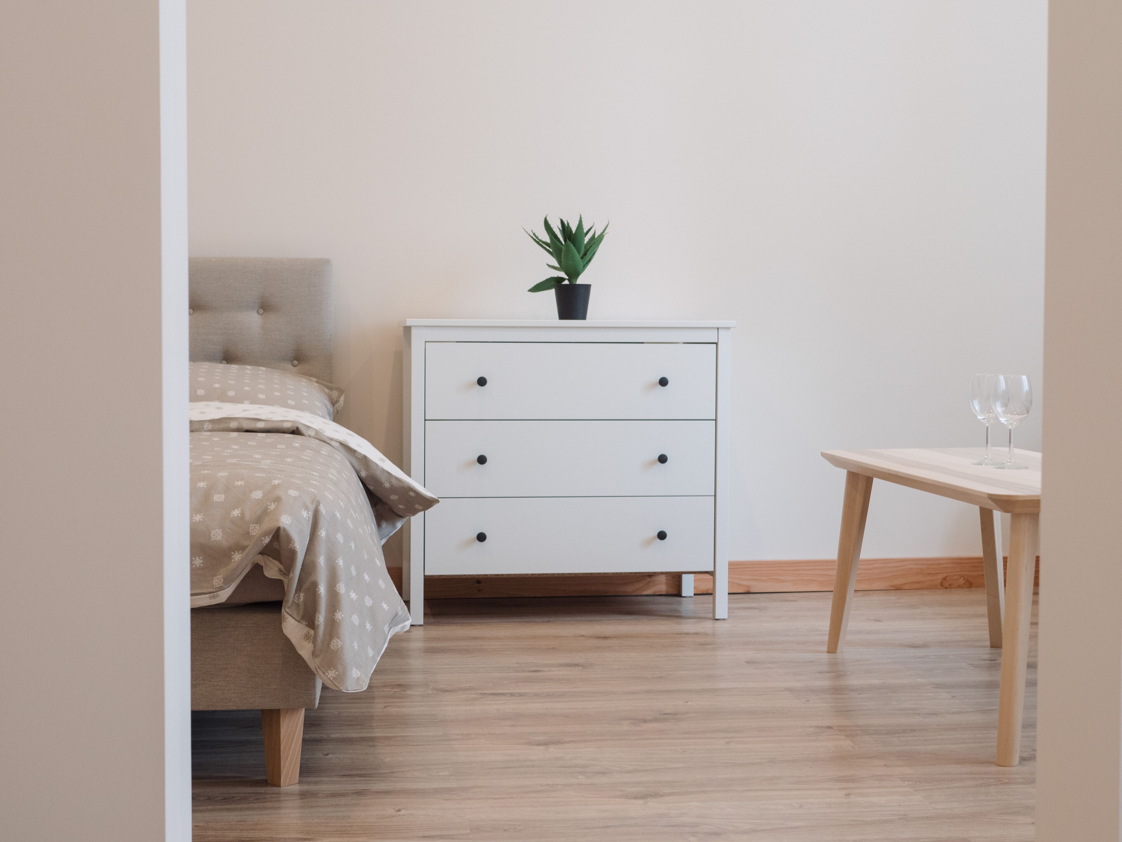 A bedroom with a bed, a nightstand, a small table, and a potted plant.
