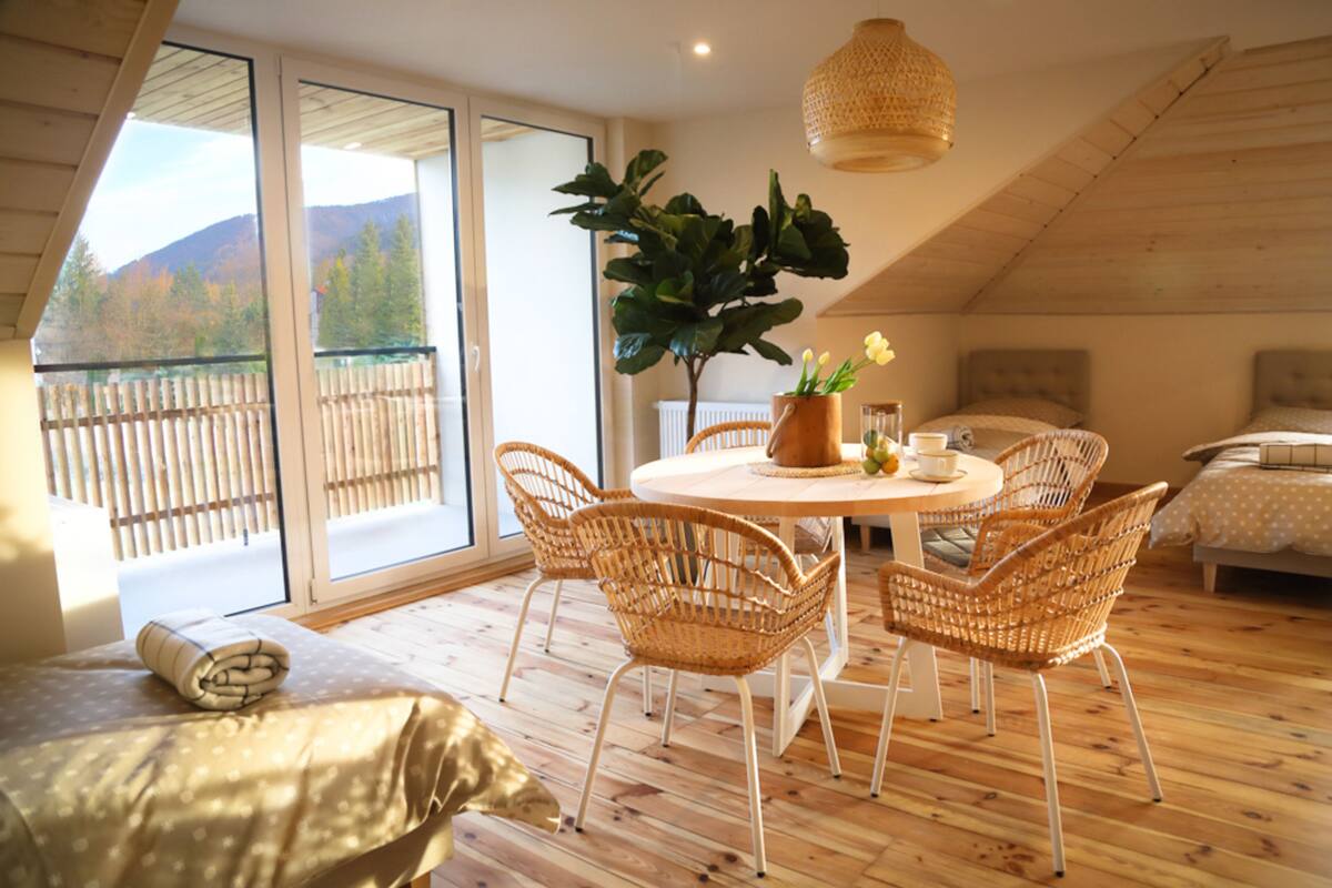 A cozy room with a wooden floor, a round table with chairs, a large potted plant, and a bed with a headboard.