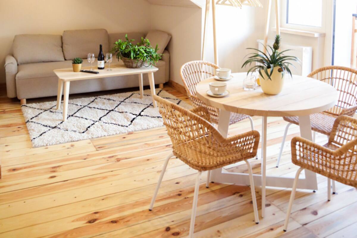 A cozy living room with a wooden floor, a round dining table with wicker chairs, a beige sofa, and a small table with a plant and wine glasses.