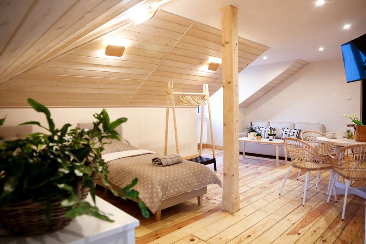 A cozy attic bedroom with a bed, a wooden ceiling, a TV, and a dining area with wicker chairs.