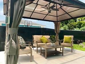 An outdoor patio with two beige cushioned chairs, a wooden table, and a ceiling fan. - Vacation in  the Comfort of Home at LiShay’s Cottage (Nassau)