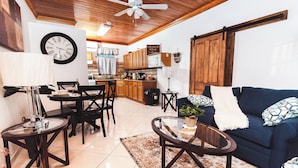 Uma sala de estar aconchegante com sofá, mesa de centro e área de refeições. Um relógio está fixado na parede, e há uma cozinha ao fundo.