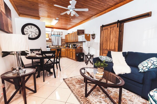 Una sala de estar acogedora con un sofá, una mesa de centro y una zona de comedor. Un reloj está colgado en la pared, y se ve una cocina al fondo.