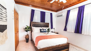 Quarto com cama de madeira, roupa de cama branca, almofada com a palavra 'RELAX', uma planta em vaso e cortinas roxas.