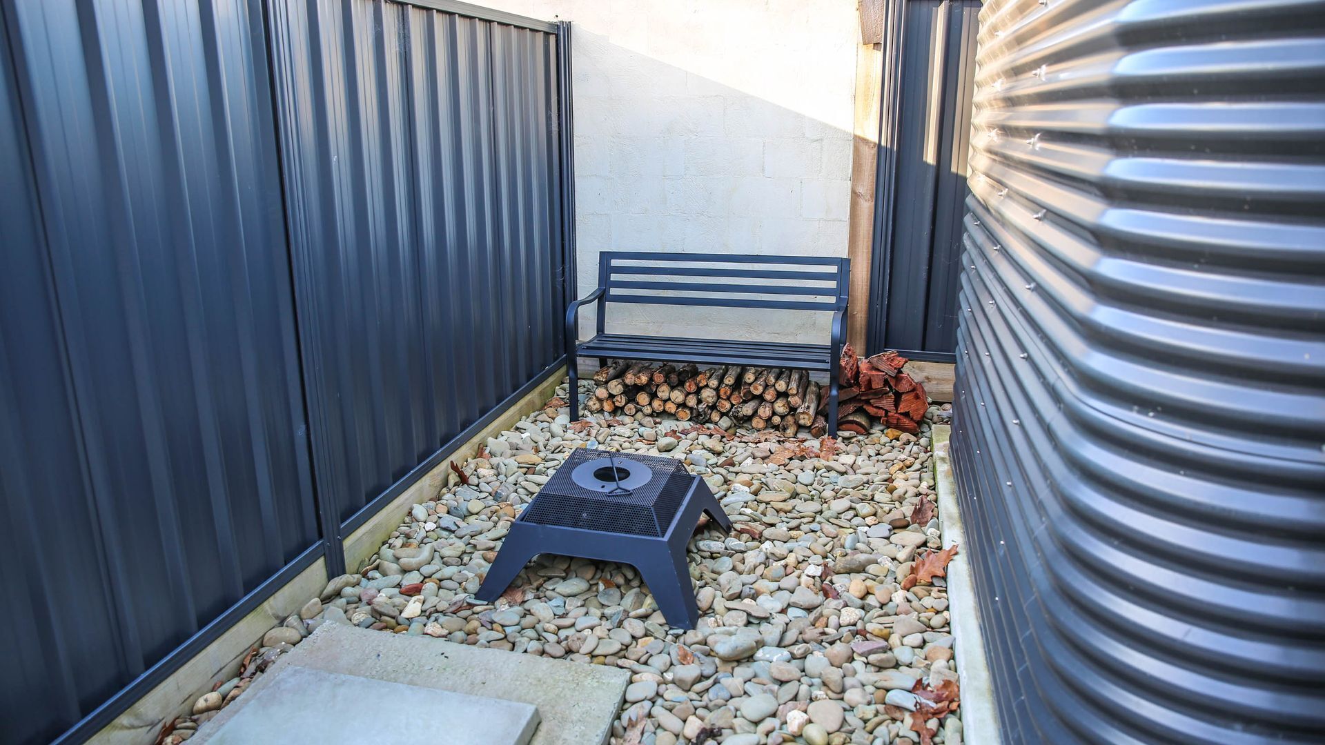 A small outdoor area with a metal bench, a small table, and a pile of firewood.
