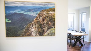 Un comedor con una mesa y sillas, un cuadro de paisaje grande en la pared, y vistas al exterior.