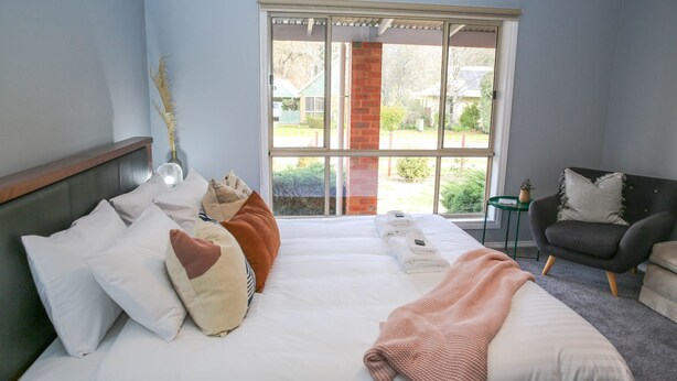 Een slaapkamer met een bed, kussens, een stoel en een raam met uitzicht op een tuin en een huis.