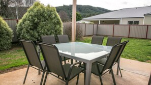 A patio with a glass-top table and six chairs, surrounded by a metal fence and a large coniferous tree. - SHINE BRIGHT - Spacious family friendly group living in Bright (Bright)