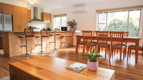 A modern kitchen with wooden cabinets, a stainless steel refrigerator, and a built-in stove. There are four bar stools and a dining table with six chairs. A potted plant is on the table, and the room has large windows. - SHINE BRIGHT - Spacious family friendly group living in Bright (Bright)