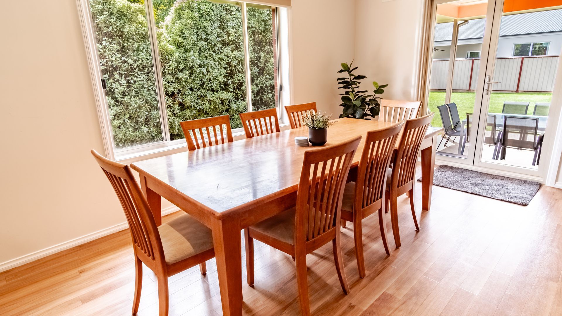 A dining area with a wooden table and chairs, a potted plant, and a view of a garden through large windows.