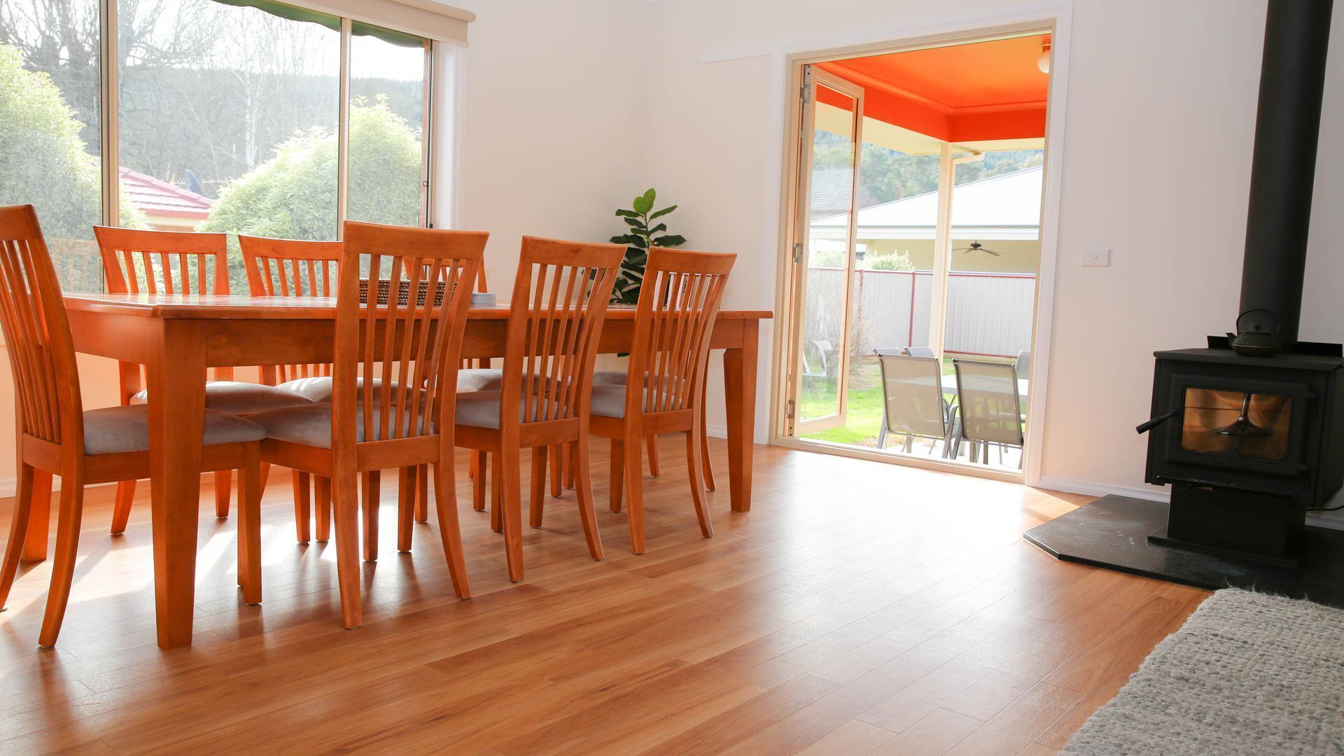 Un coin repas avec une table et des chaises en bois, un poêle en bois noir et une vue sur l’extérieur grâce à de grandes fenêtres.