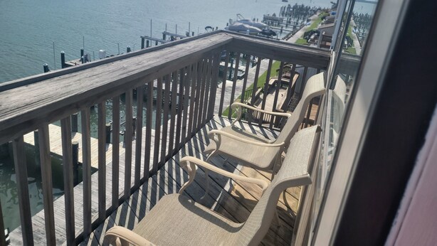 Un balcón con barandas de madera y sillones de descanso con vista a un puerto deportivo con barcos y una costa lejana.