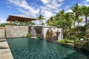 A pool with a waterfall feature, surrounded by palm trees and a stone wall. - Champion Ridge #35: Paradisiac Home w/Infinity Pool, Hot Tub & Tropical Feeling (Kamuela)