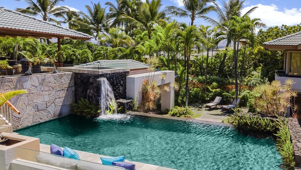 Uma piscina com cachoeira, rodeada por vegetação tropical exuberante e palmeiras.