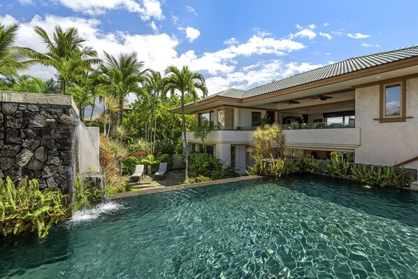 A pool area with a stone waterfall feature, surrounded by lush greenery and palm trees. - Champion Ridge #35: Paradisiac Home w/Infinity Pool, Hot Tub & Tropical Feeling (Kamuela)