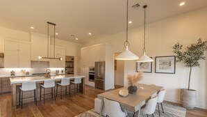 A modern kitchen with a central island, bar stools, pendant lighting, and a dining area with a table and chairs. - The 19th Hole at The Ledges (St. George)