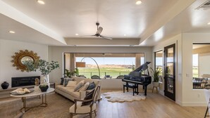A spacious living room with a fireplace, a piano, a large window with a view of a field, and a ceiling fan. - Taking Flight (St. George)