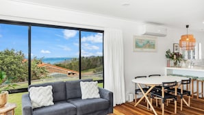 Una sala de estar moderna con un gran ventanal que ofrece una vista panorámica, un sofá gris con cojines, una mesa de comedor con sillas y una zona de cocina con una lámpara colgante.