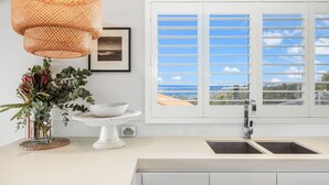 Una cocina con encimera blanca, un fregadero, una mesa blanca con base circular y un cuenco, una lámpara de mimbre y una ventana con contraventanas blancas.