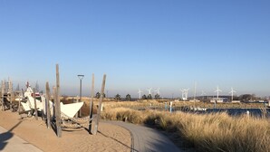 A coastal path with wind turbines in the background, a sandy area with wooden structures, and a concrete tower.