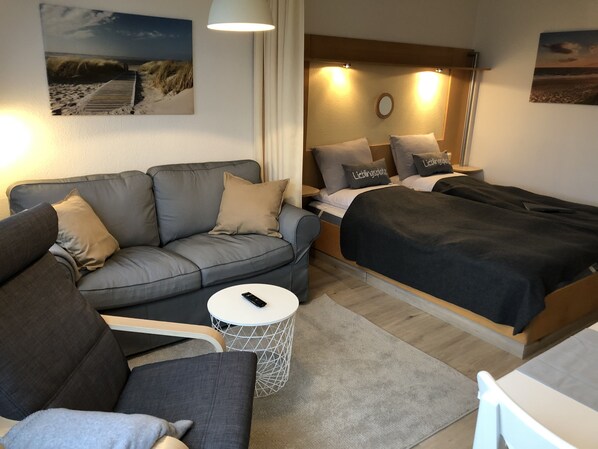 Une chambre moderne avec un canapé gris, une petite table ronde sur laquelle se trouve une télécommande, un lit recouvert d’une couverture foncée et une lampe fixée au mur.