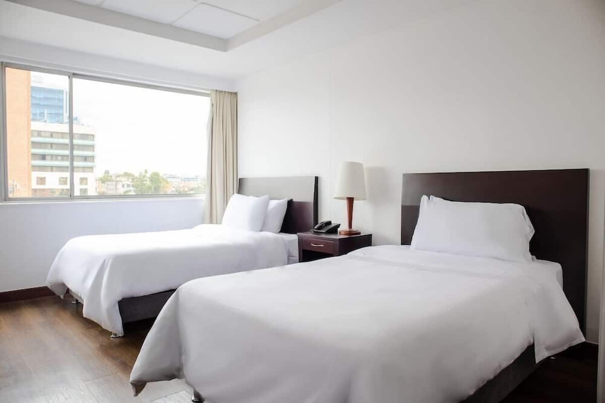 Habitación de hotel con dos camas, una mesita de noche con lámpara y un ventanal con vistas a la ciudad.
