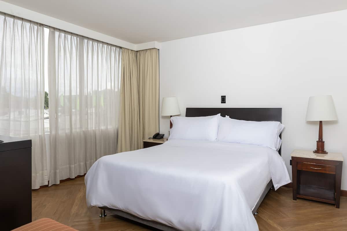 Una cama bien hecha con sábanas blancas, una mesita de noche con una lámpara y una ventana con cortinas translúcidas.