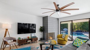 A modern living room with a flat-screen TV, a ceiling fan, a fireplace, and a view of a swimming pool. - Villa Del Sol – Modern Luxury on the Fairway (Palm Springs)