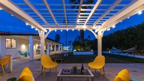 An outdoor patio with a pergola, yellow chairs, a table with a book and a bottle, and a pool area in the background. - Villa Del Sol – Modern Luxury on the Fairway (Palm Springs)