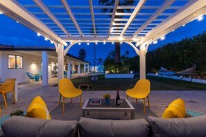 An outdoor patio with a pergola, yellow chairs, a table with a book and a bottle, and a pool area in the background.
