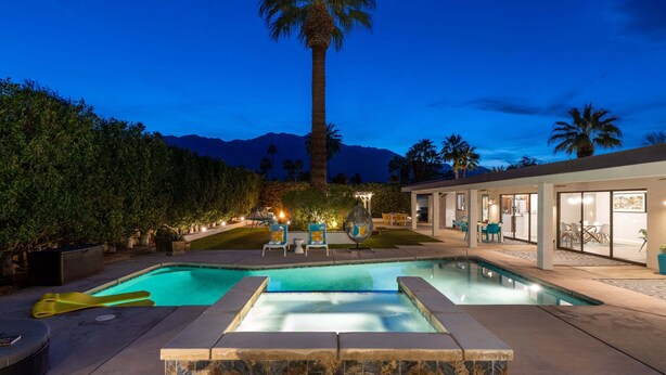 A swimming pool with lounge chairs, a palm tree, and a modern house with glass doors. - Villa Del Sol – Modern Luxury on the Fairway (Palm Springs)