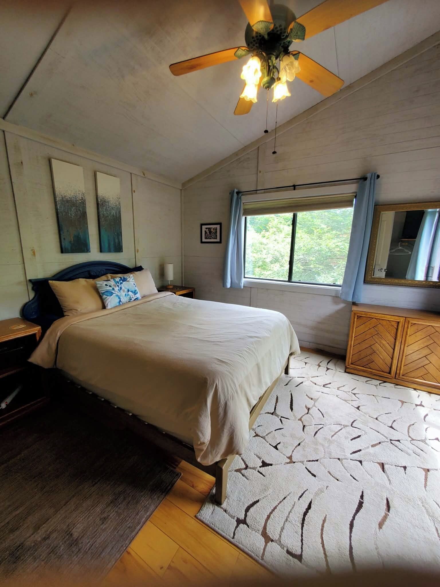 Ein Schlafzimmer mit einem Bett, einem Deckenventilator, einem Fenster mit Vorhängen, einem Holzschrank und einem Teppich.