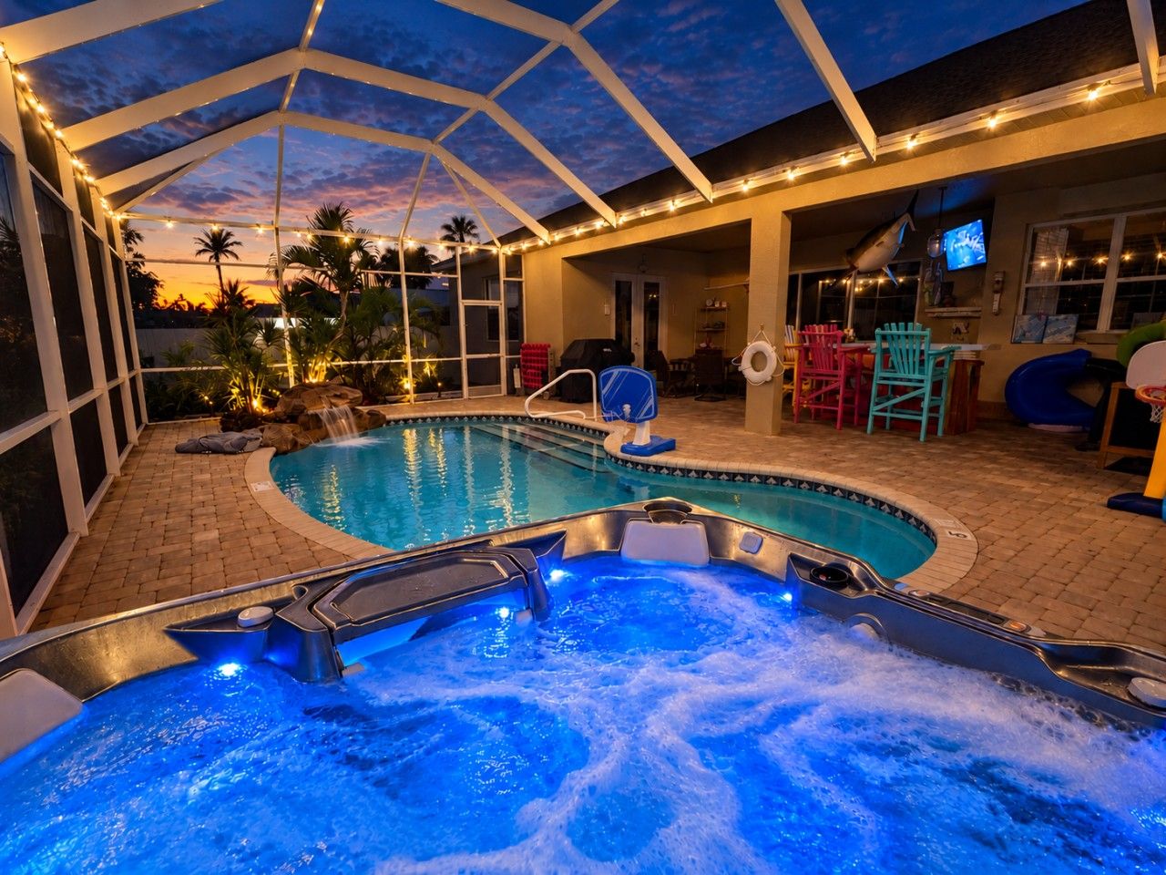 A swimming pool with a hot tub, surrounded by a covered patio area with chairs and a television.