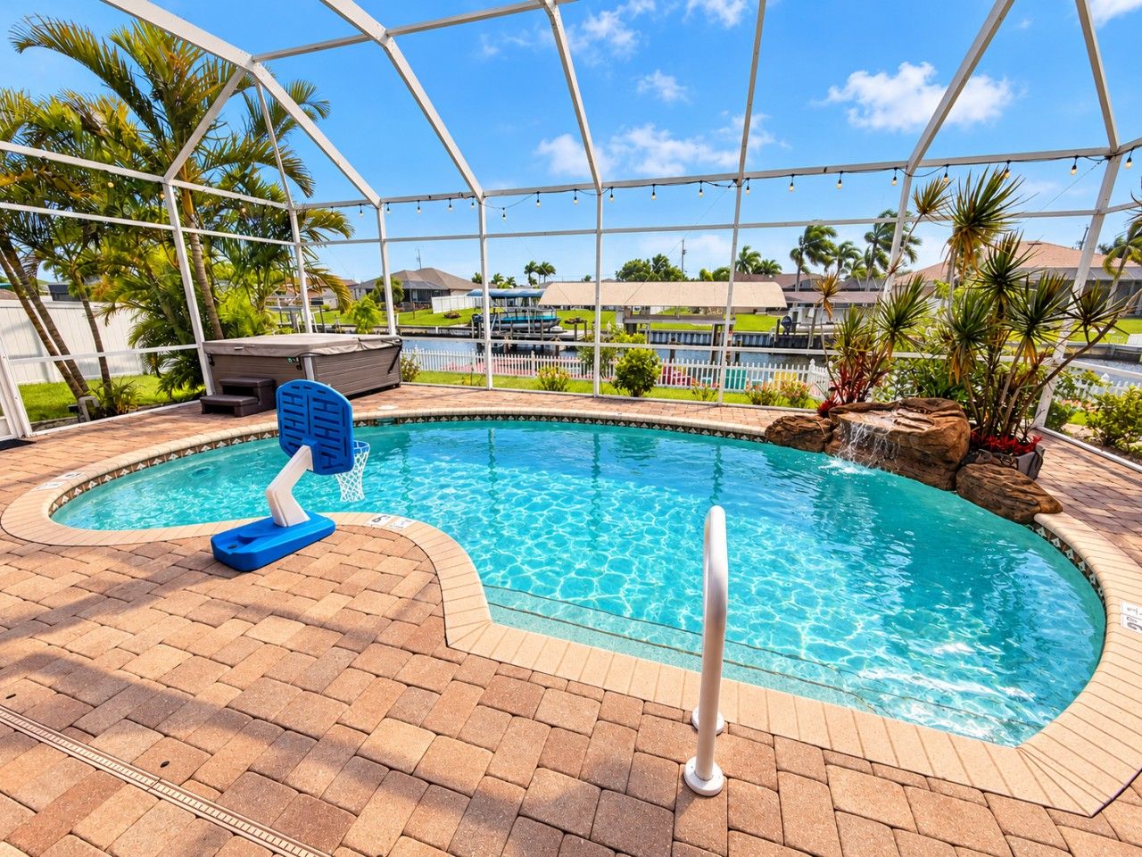 Una piscina con una característica de agua azul cristalina, rodeada por un patio de ladrillo y un área cubierta con estructura metálica y luces colgantes.