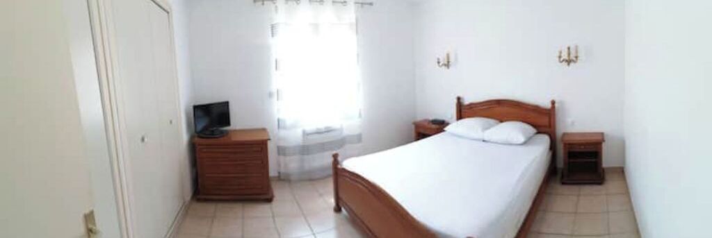 Ein Schlafzimmer mit Bett, Nachttisch, Kommode, Fernseher und einem Fenster mit Vorhängen.