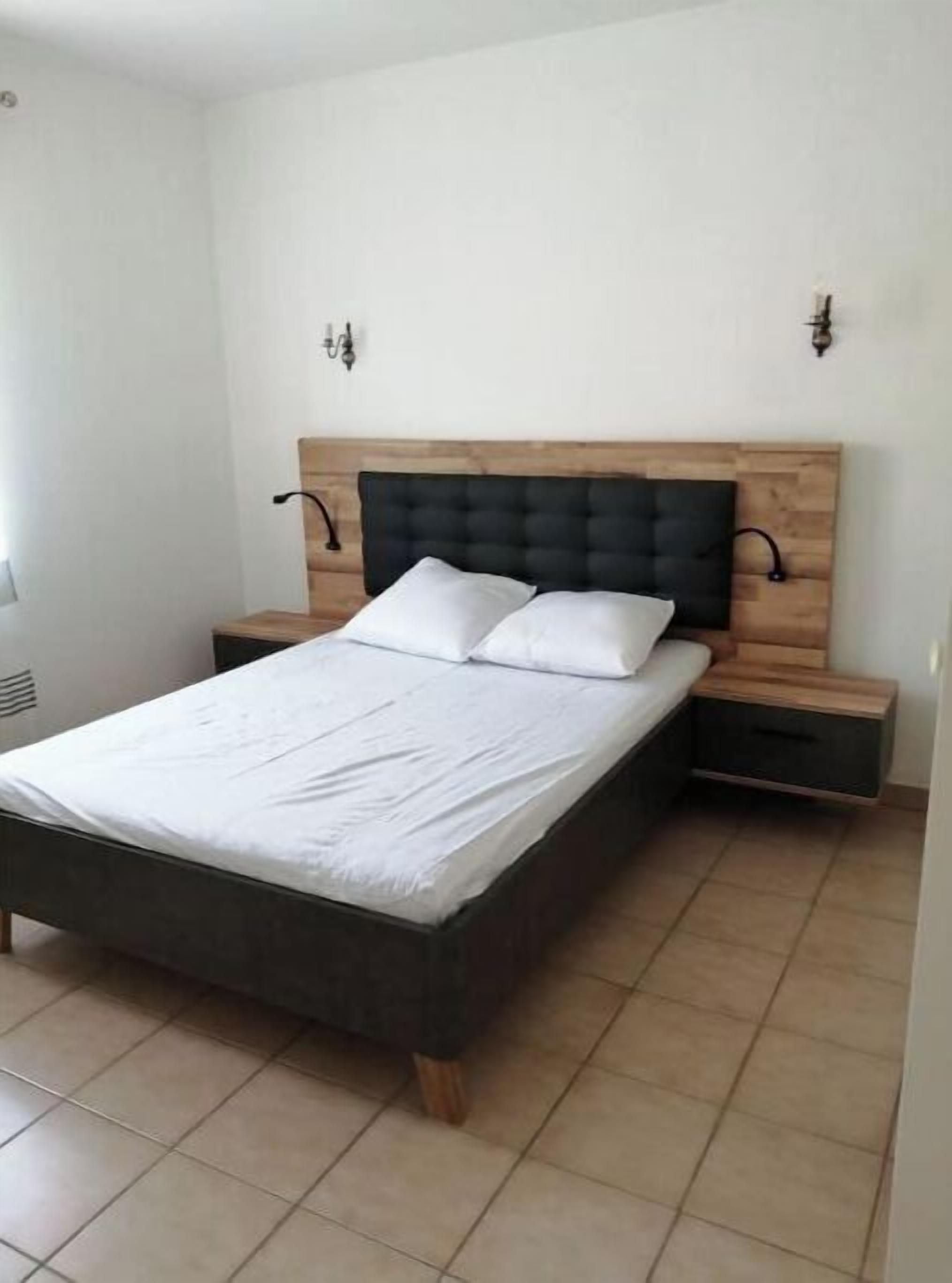 A bedroom with a wooden headboard, a bed with white linens, two bedside tables, and a wall-mounted light fixture.