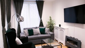 A modern living room with a grey sofa, black armchairs, a glass coffee table, a floor lamp, a TV, and a fireplace. - Guthrum Place 3bedroom Luxury Apt Royal Wharf Dock (London)