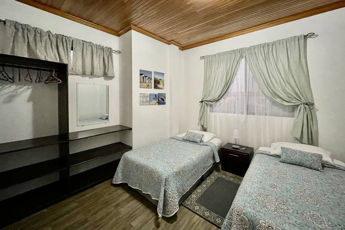 A bedroom with two beds, a wooden ceiling, a window with curtains, a mirror, and a wall with pictures.