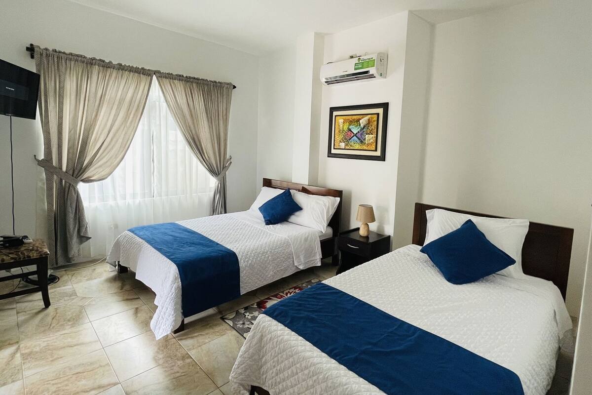 A hotel room with two beds, a TV, a window with curtains, a wall-mounted air conditioner, and a framed picture.