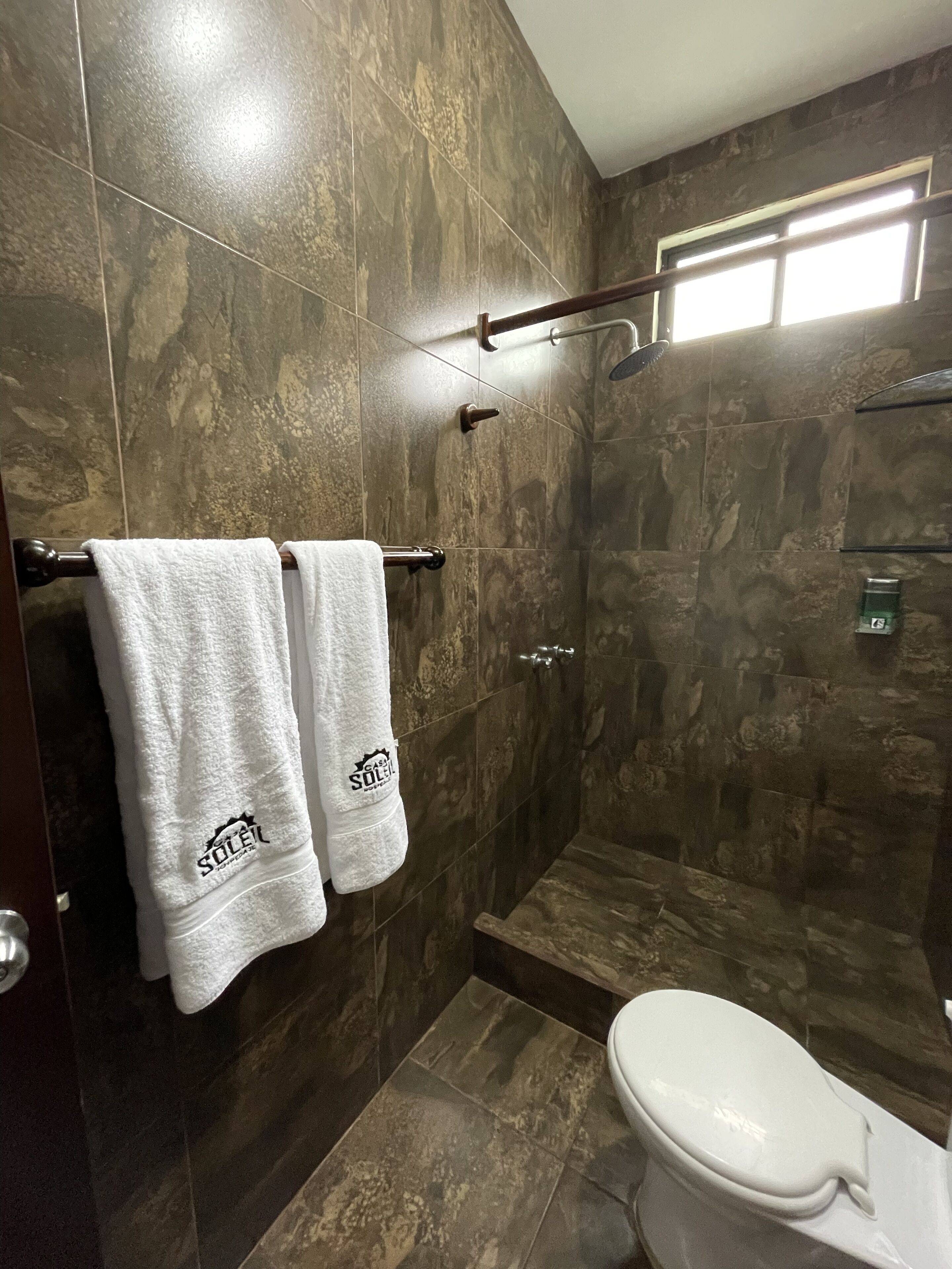 Un baño con baldosas de mármol oscuro, un inodoro blanco y dos toallas colgadas en un perchero.