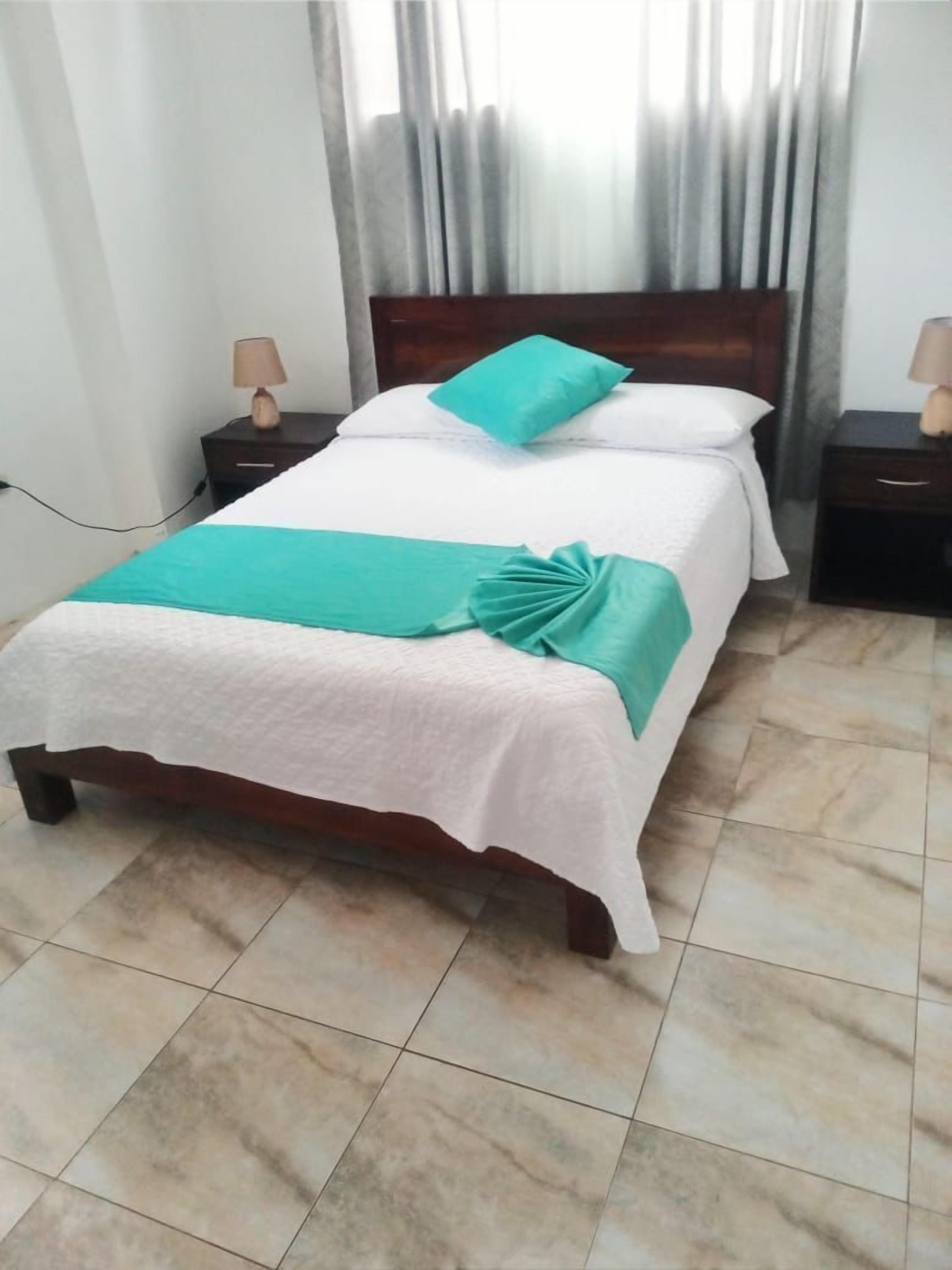 A neatly made bed with a white and teal bedspread, a teal pillow, and a folded teal blanket. There are two bedside tables with lamps, and the room has tiled flooring.