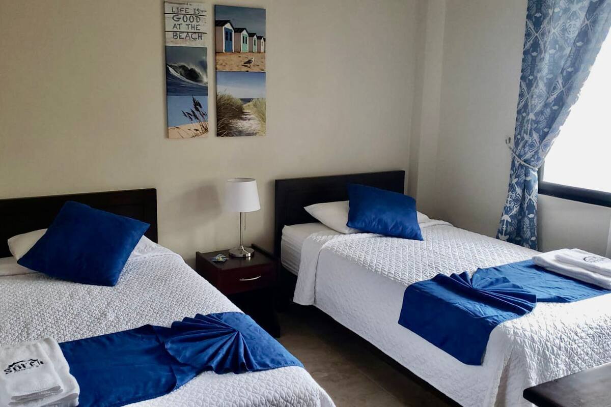 A hotel room with two beds, each with blue bedding and pillows, a small bedside table with a lamp, and a wall with a beach-themed artwork.