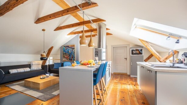 A modern loft apartment with exposed wooden beams, a central island with bar stools, a kitchen area, and a living space with a sofa and coffee table.