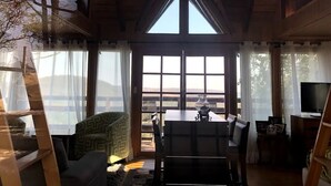 Un intérieur de chalet chaleureux avec une grande fenêtre, des poutres en bois, un coin repas et un téléviseur.